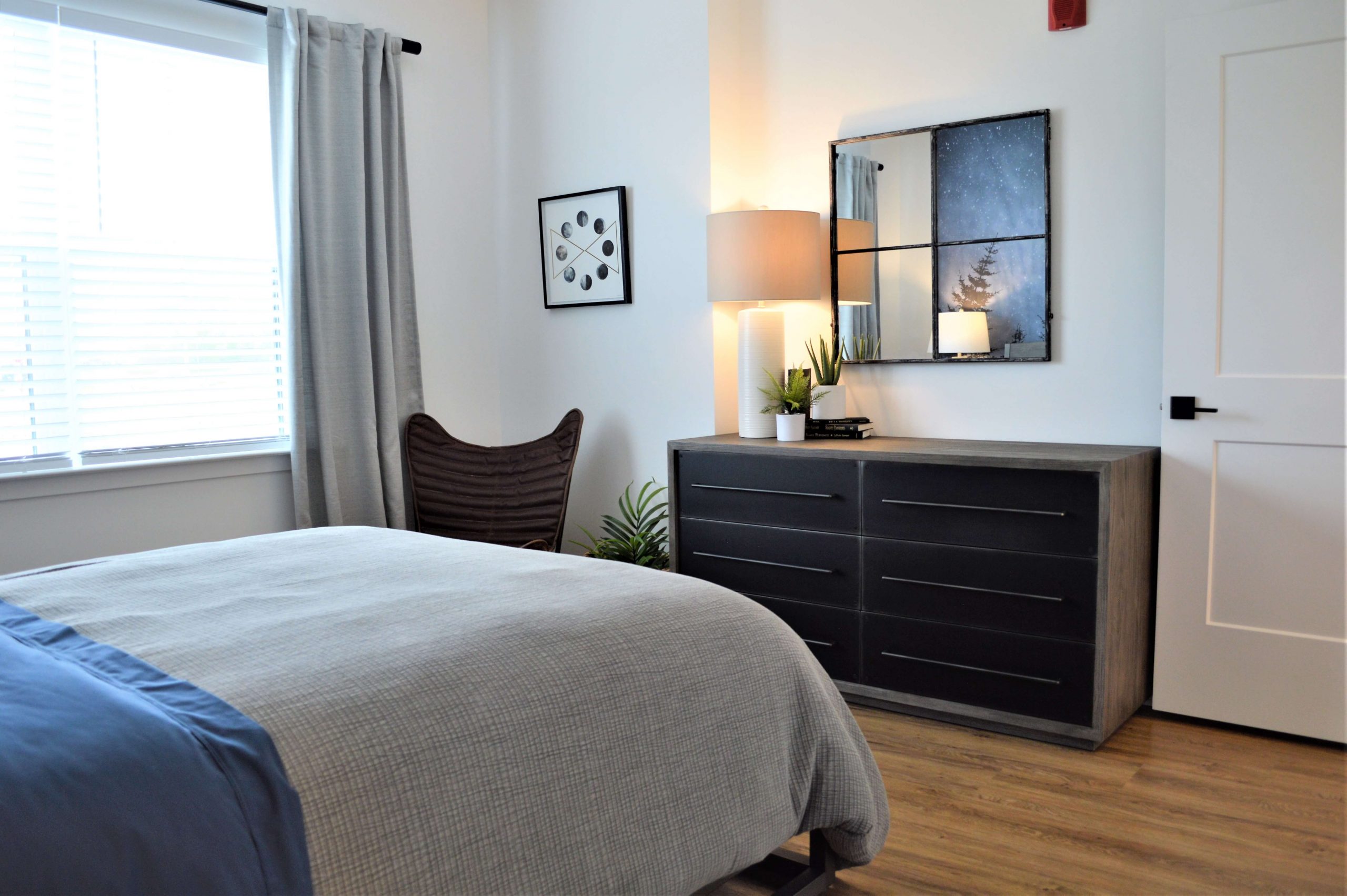 Bedroom with black dresser, mirror, table lamp, moon phase art, and bed with gray linens near a large window
