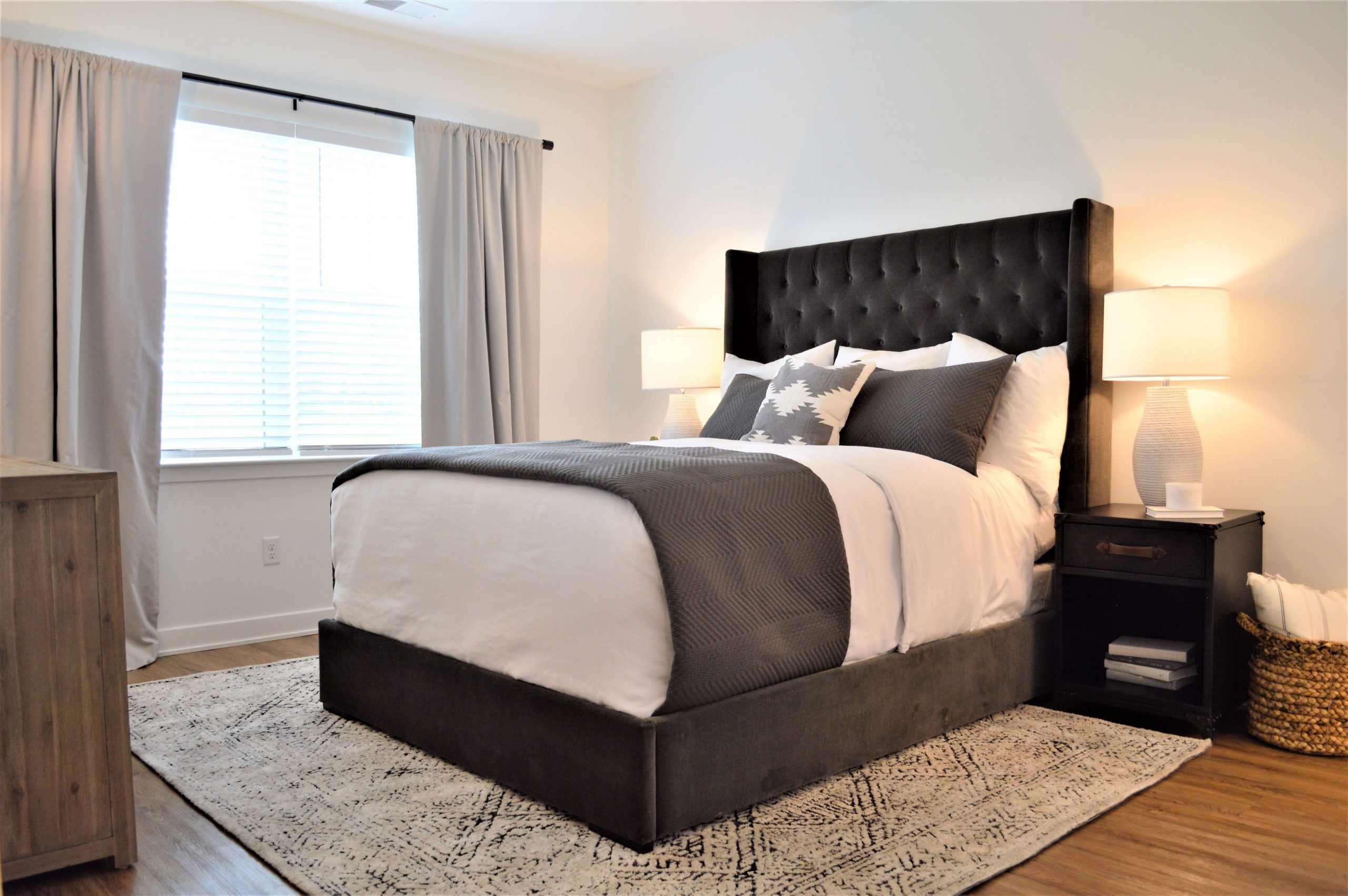 Bedroom with tufted upholstered headboard, gray bedding, nightstands with lamps, and a large window with curtains