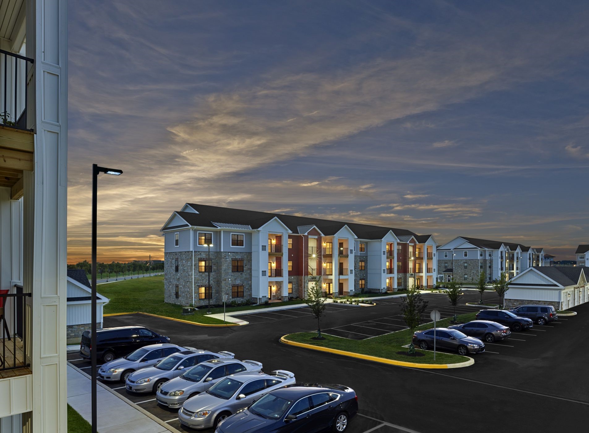 Apartment building exterior at dusk with illuminated windows and a parking lot filled with cars in the foreground.
