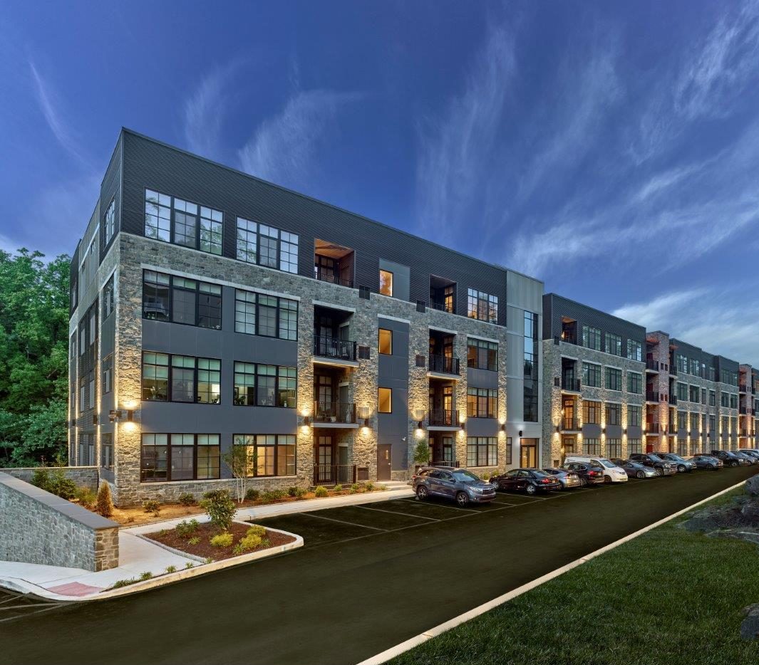 Multi-story apartment building at The Falls with stone and metal facade, illuminated at dusk with cars parked along the drive.
