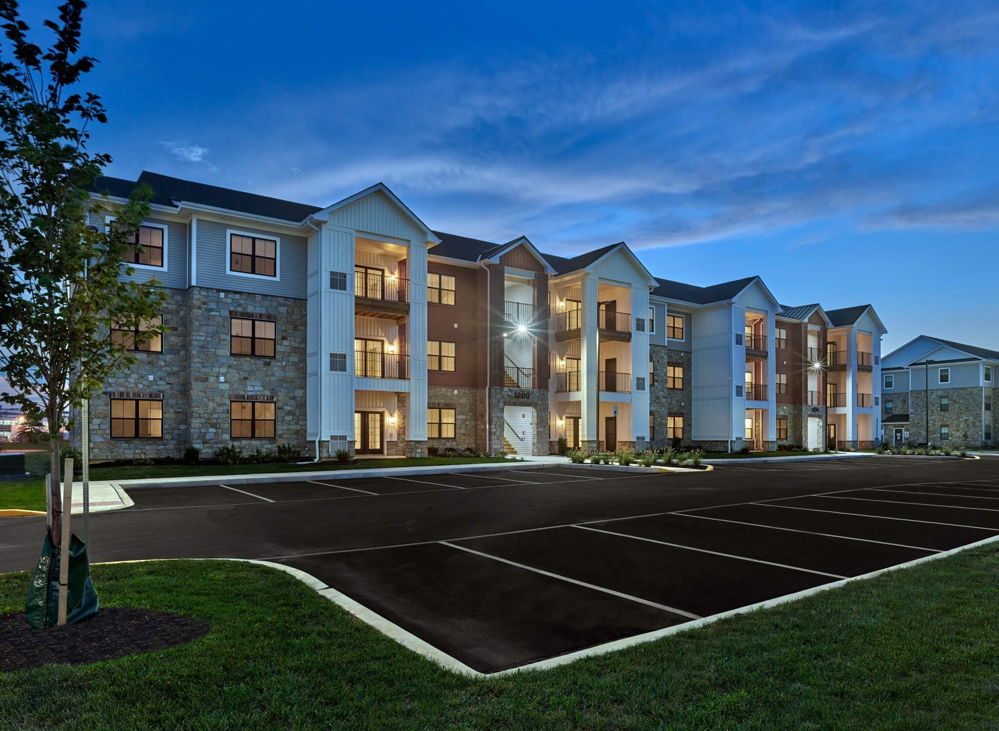 Three-story apartment building with stone and siding exterior illuminated at dusk, overlooking a paved parking lot.