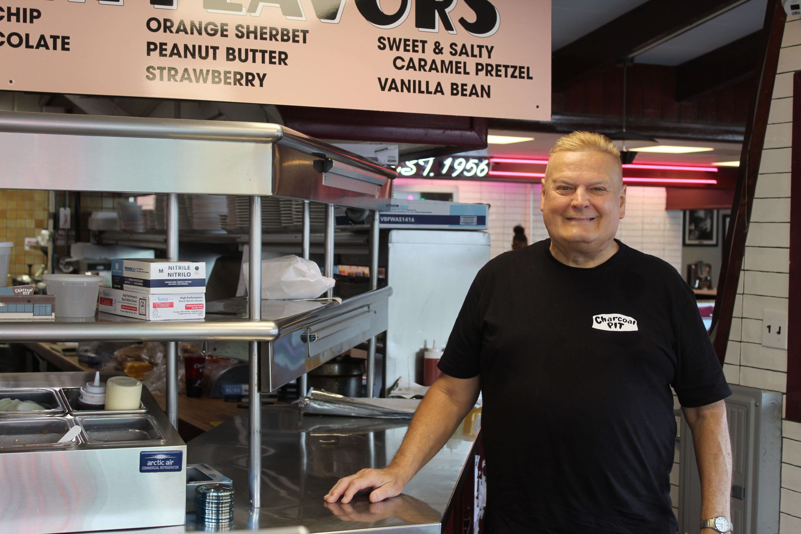 Smiling man in a Charcoal Pit shirt stands beside stainless steel kitchen counters under a hand-dipped flavors menu board.