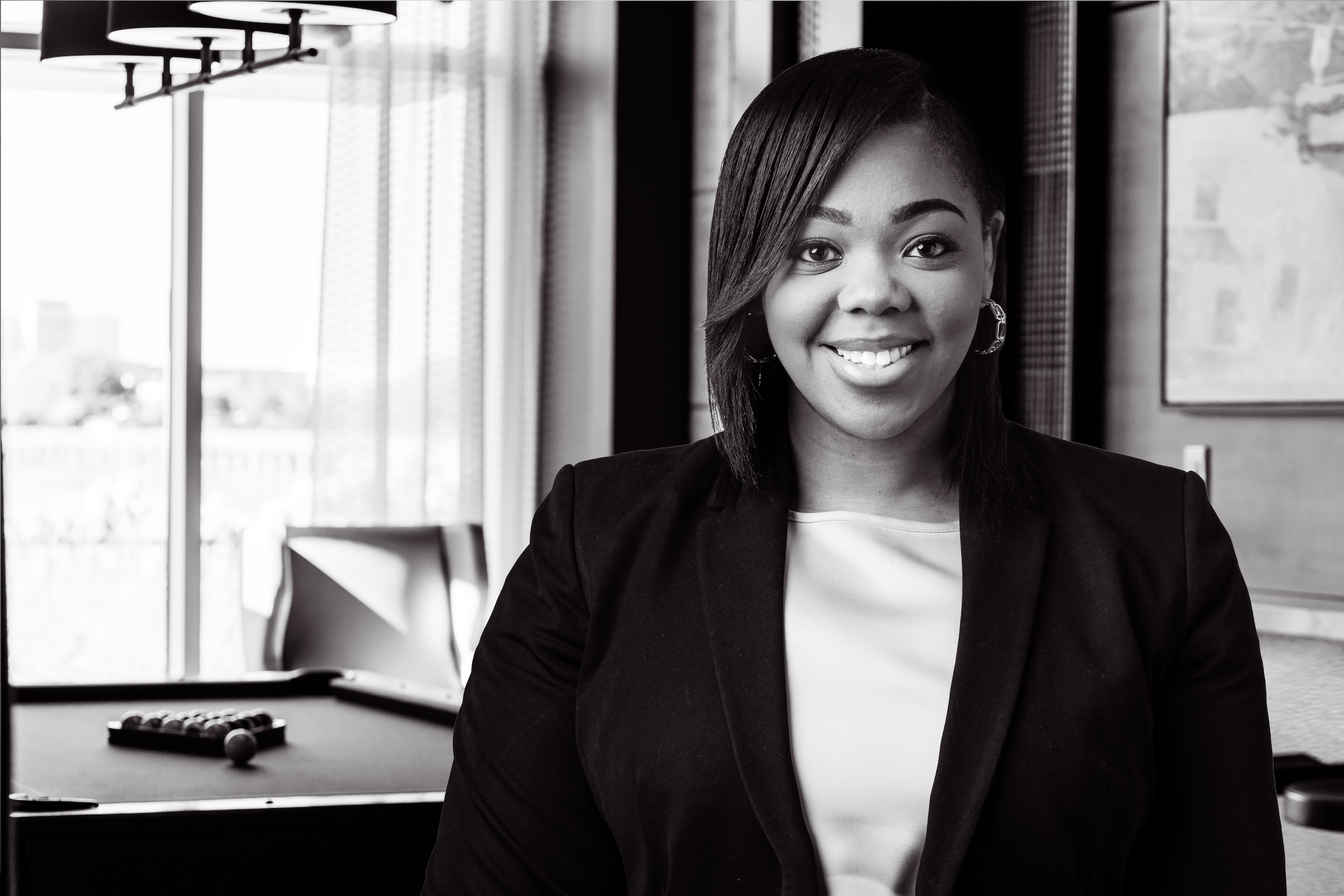 Smiling businesswoman in a blazer poses indoors near a window and pool table
