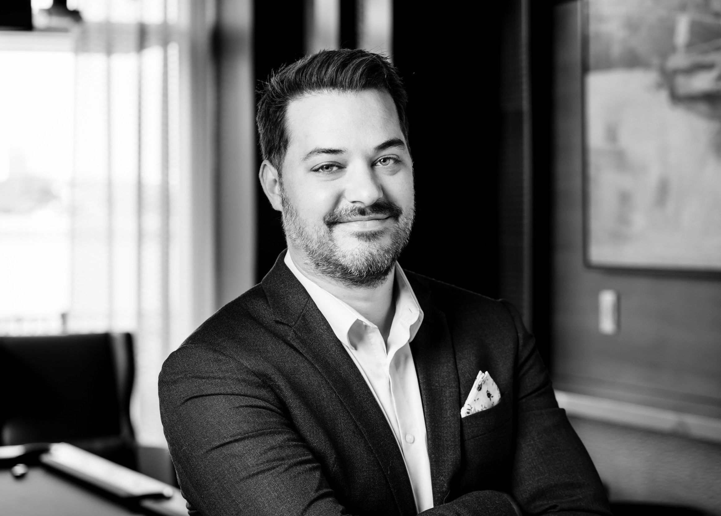 Smiling businessman in a suit with arms crossed poses for a professional portrait in an office setting.
