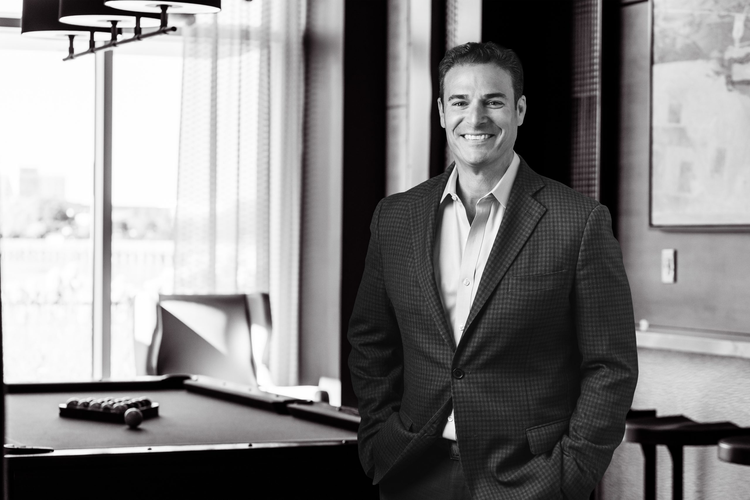 Smiling man in a checkered blazer stands near a pool table in a lounge with large windows.