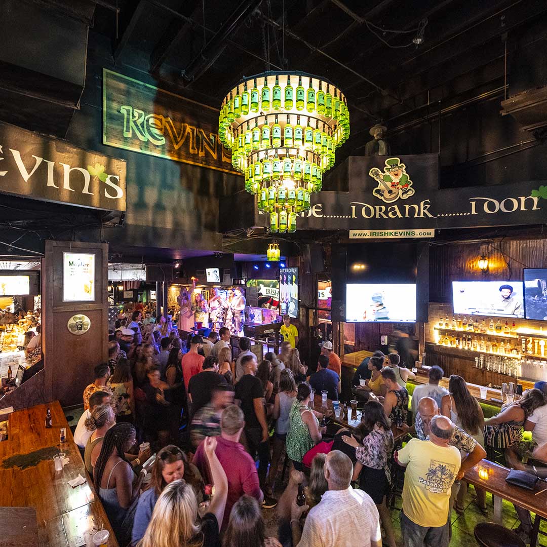 A crowded Irish Kevins bar with a bottle-cap chandelier, live music stage, and patrons filling the space beneath wall signage.