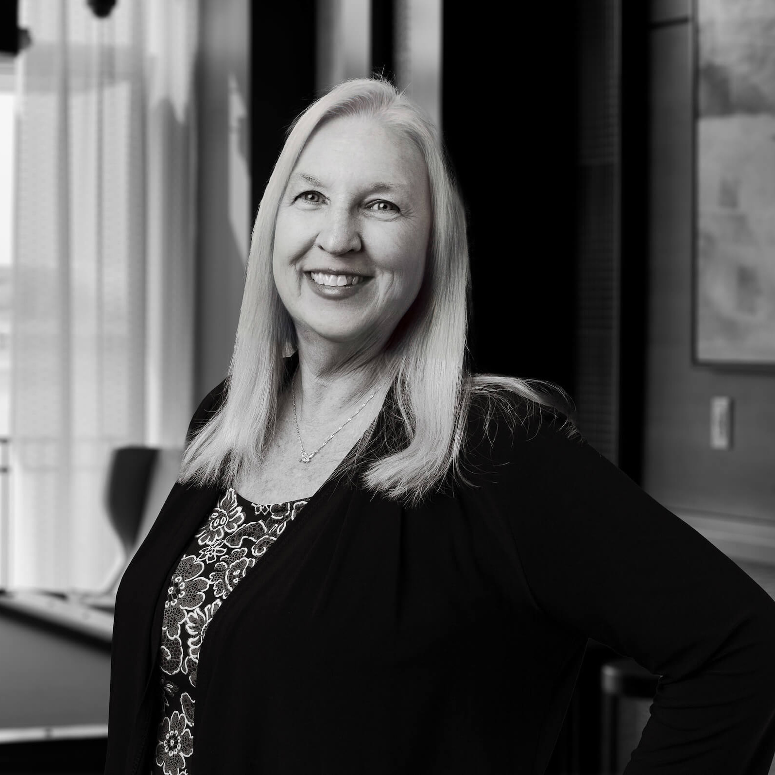Smiling woman with long blonde hair wearing a black cardigan and floral top in a black and white office portrait