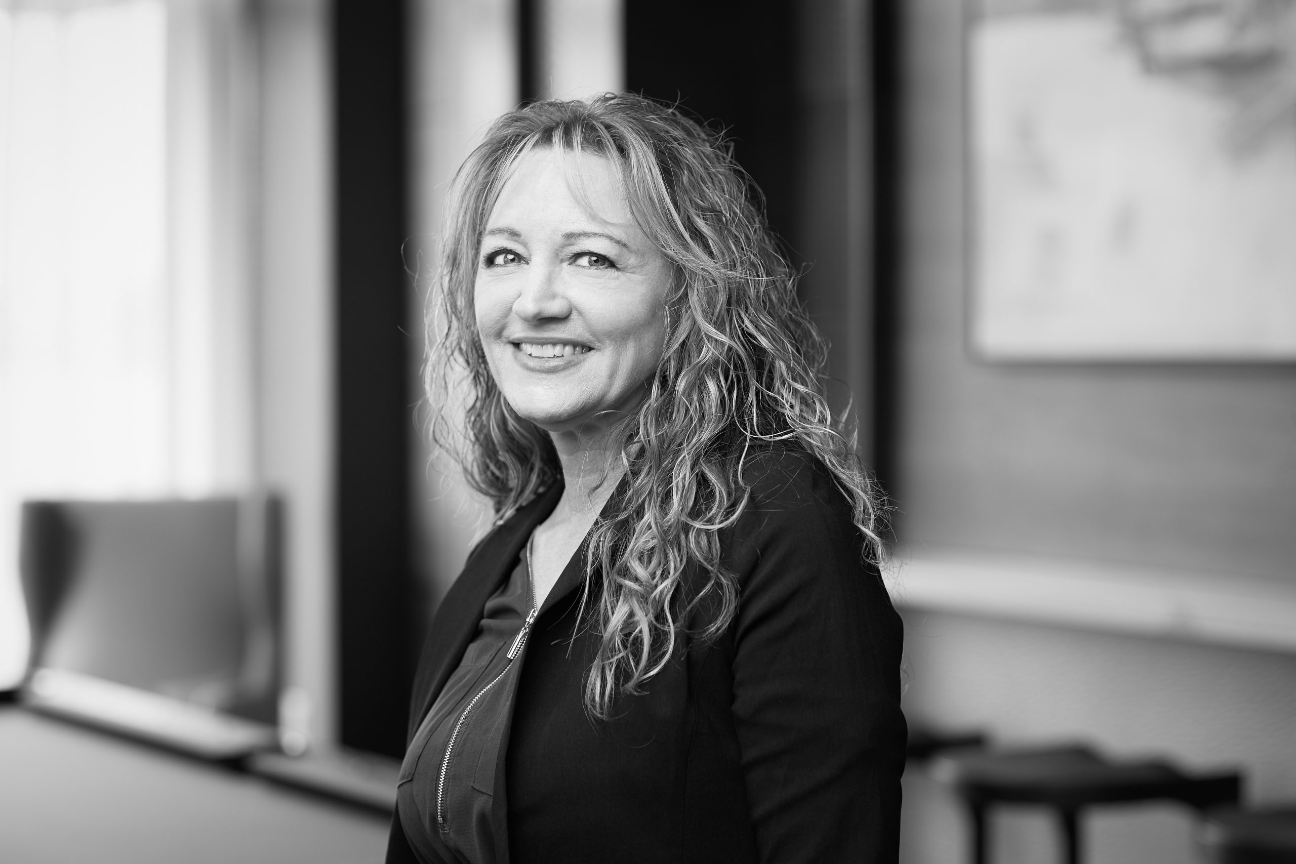 Woman with curly hair smiling in a professional office setting, wearing a black jacket.