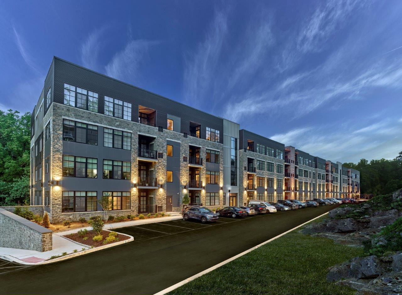 Multi-story stone and metal-paneled apartment building at The Falls with illuminated windows and balconies at dusk, with cars parked along the drive.