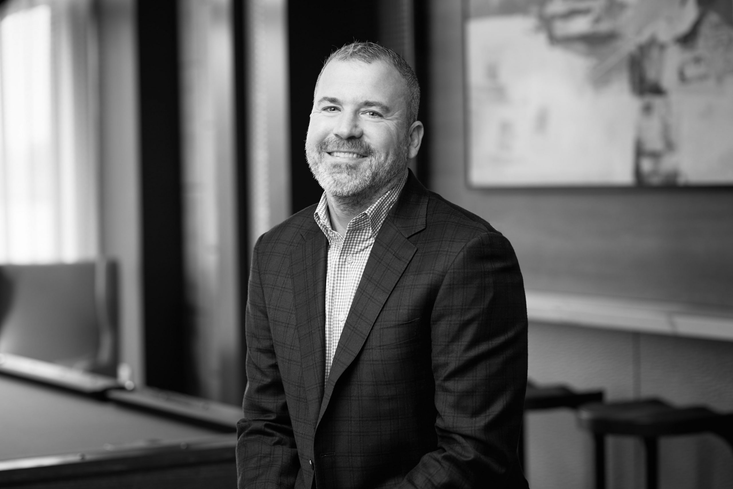 Smiling bearded man in a plaid blazer and checked shirt poses in a modern office setting.