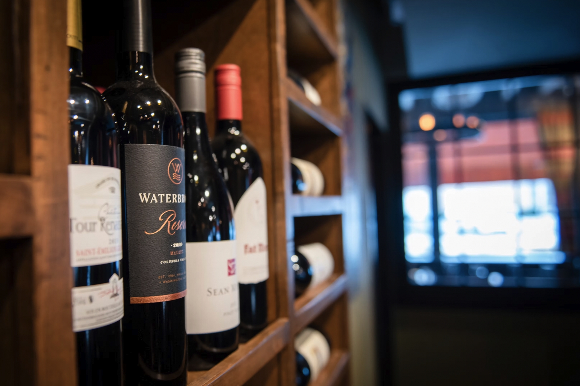 Wine bottles displayed on a wooden wall rack with a blurred bar area in the background.