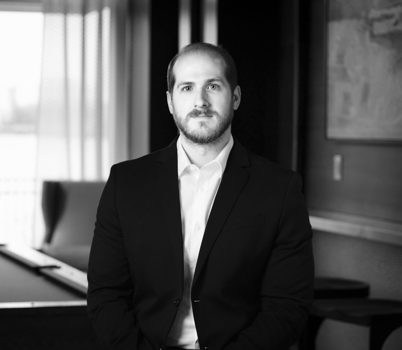 Bearded man in a dark suit and white shirt stands in an office lounge with a curtained window behind him.