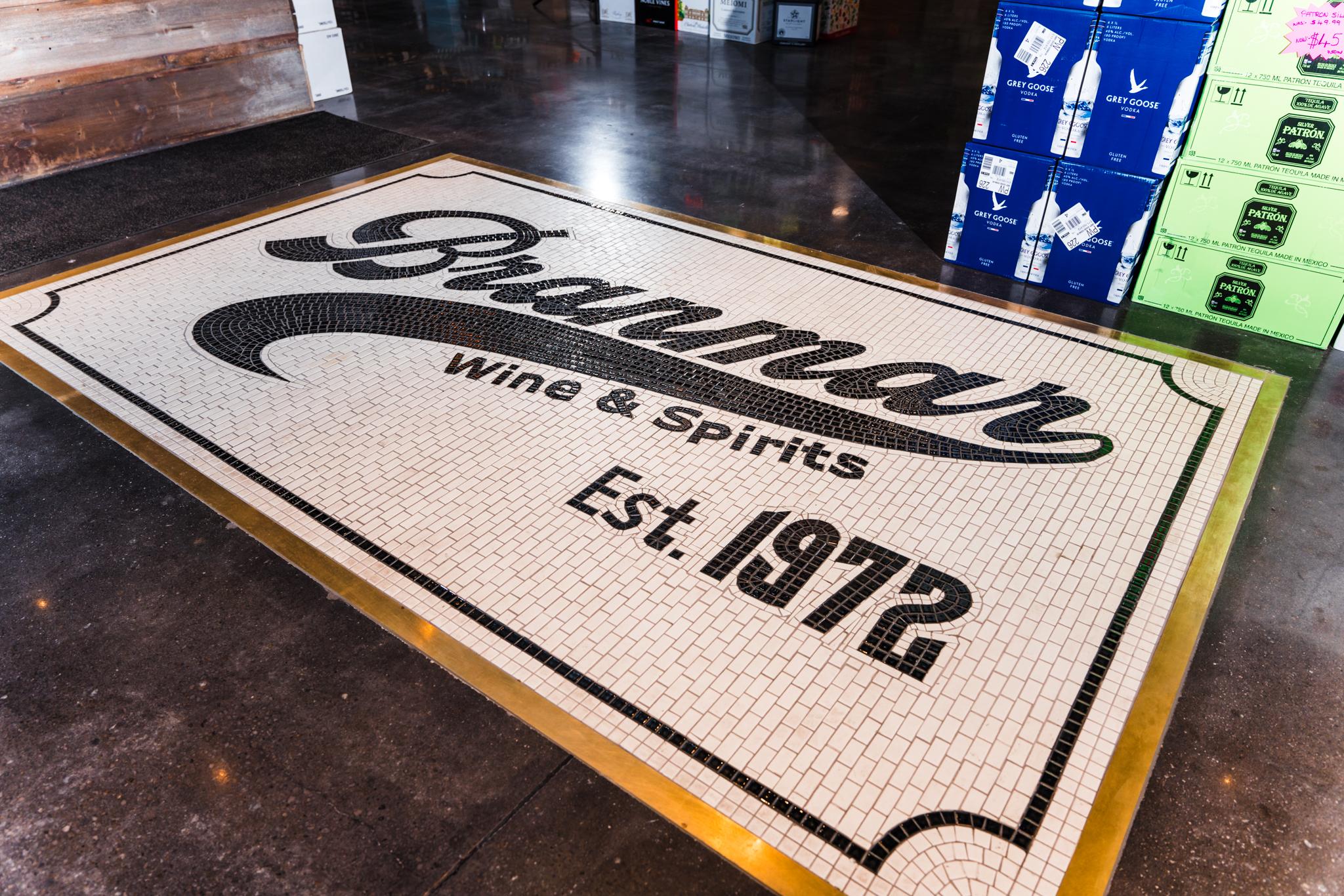 Mosaic tile floor entrance reading Branman Wine & Spirits Est. 1972, with wine and liquor cases stacked nearby.