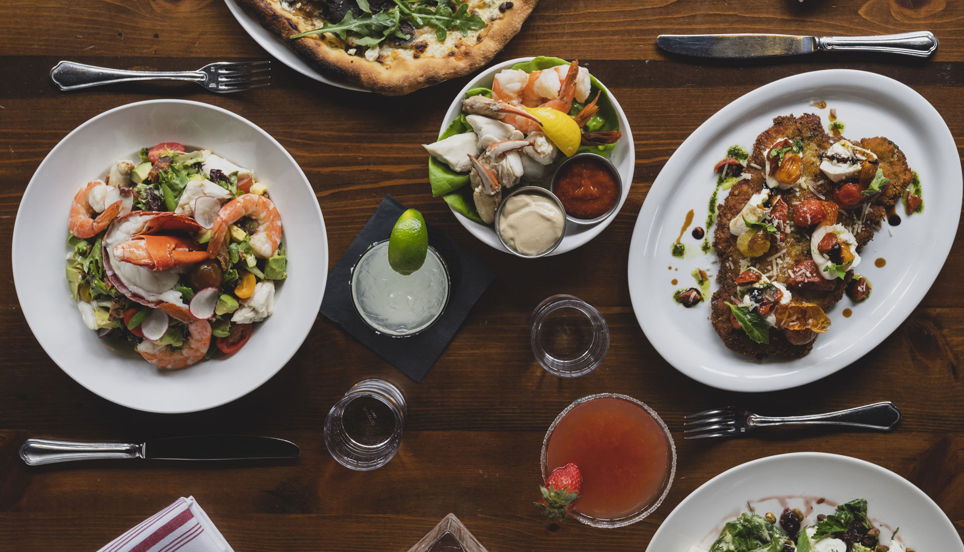 Overhead view of a wooden table set with seafood salad, chilled shellfish, pizza, entrees, and cocktails