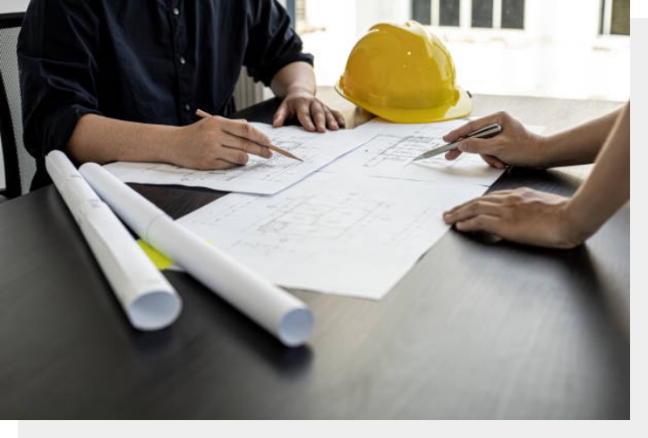 Two people review architectural blueprints at a table with a hard hat and rolled plans nearby