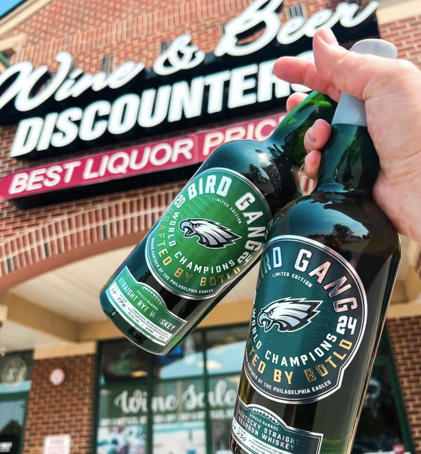 A hand holds two Bird Gang limited edition whiskey bottles outside the Wine & Beer Discounters storefront at Dove Run Wine & Spirits.