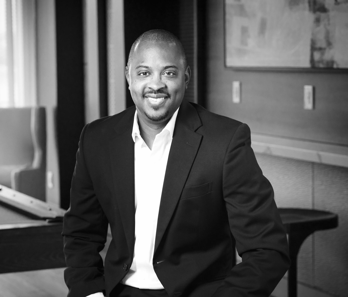 Smiling bald man with a goatee wears a black blazer over a white shirt in a modern office setting.