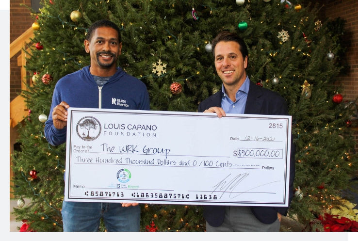 Two men hold a large ceremonial check for $300,000 from the Louis Capano Foundation to The WRK Group in front of a decorated Christmas tree.
