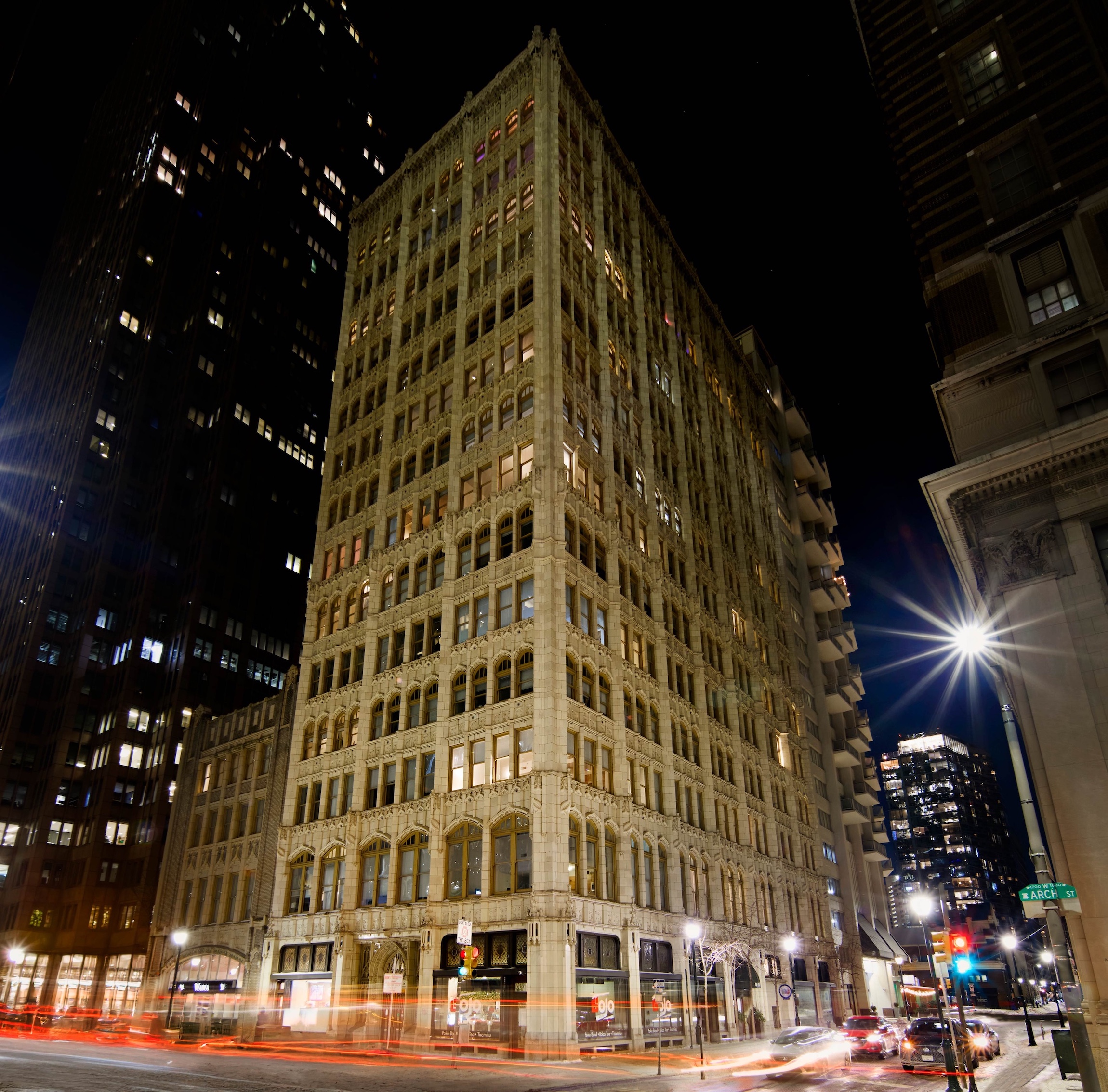 The Arch Luxury Apartments building rises above a city street corner illuminated at night with light trails from passing traffic.