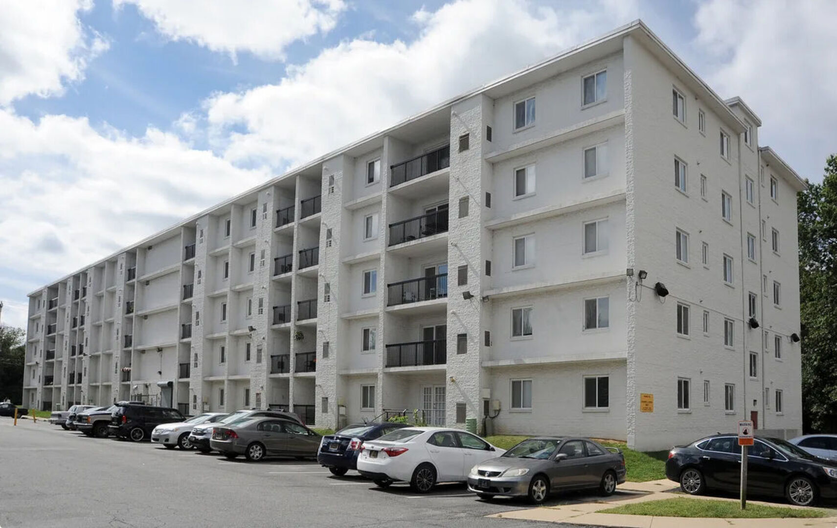 Five-story white apartment building with balconies and a parking lot filled with resident vehicles.