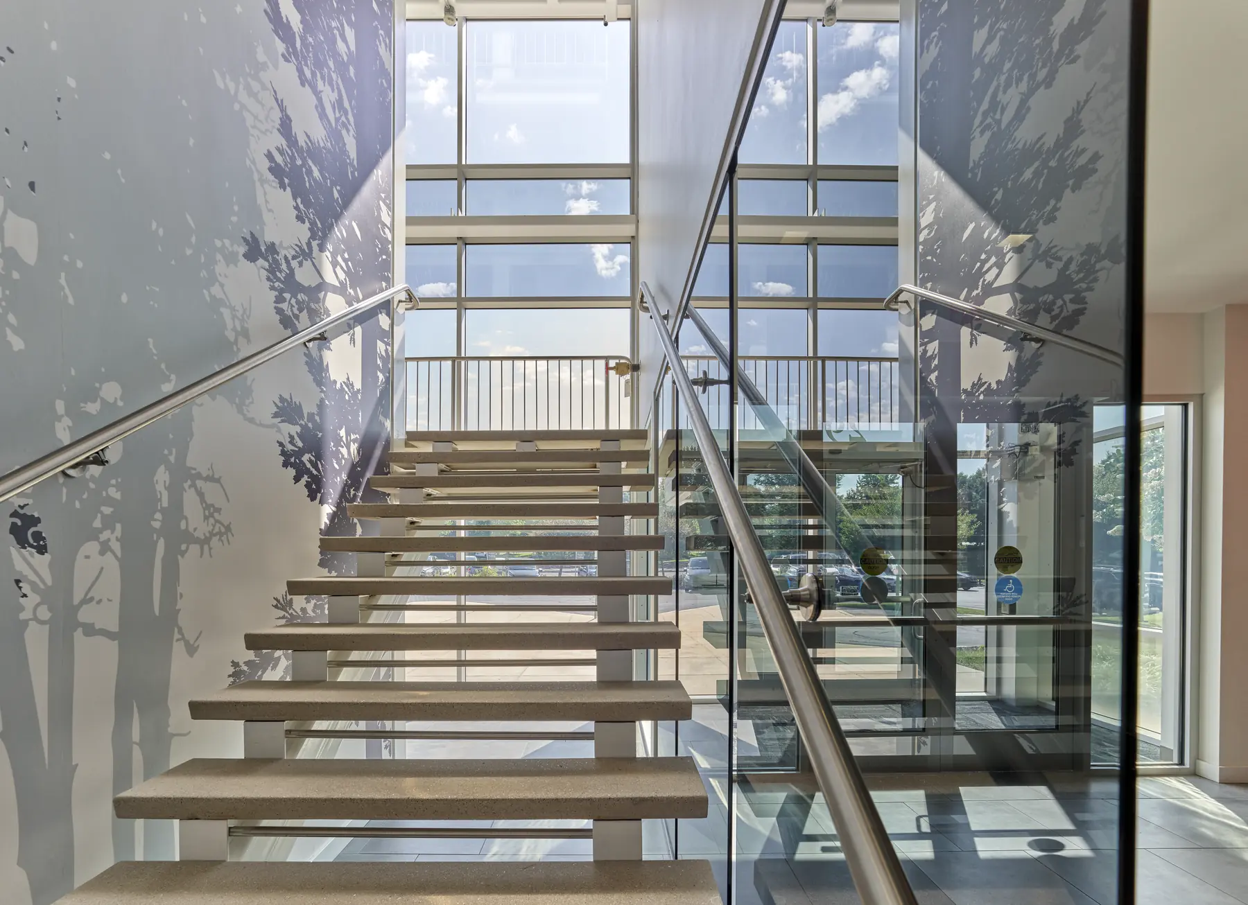 Floating staircase with glass railings and tree-silhouette wall art beside floor-to-ceiling windows in an office building lobby.