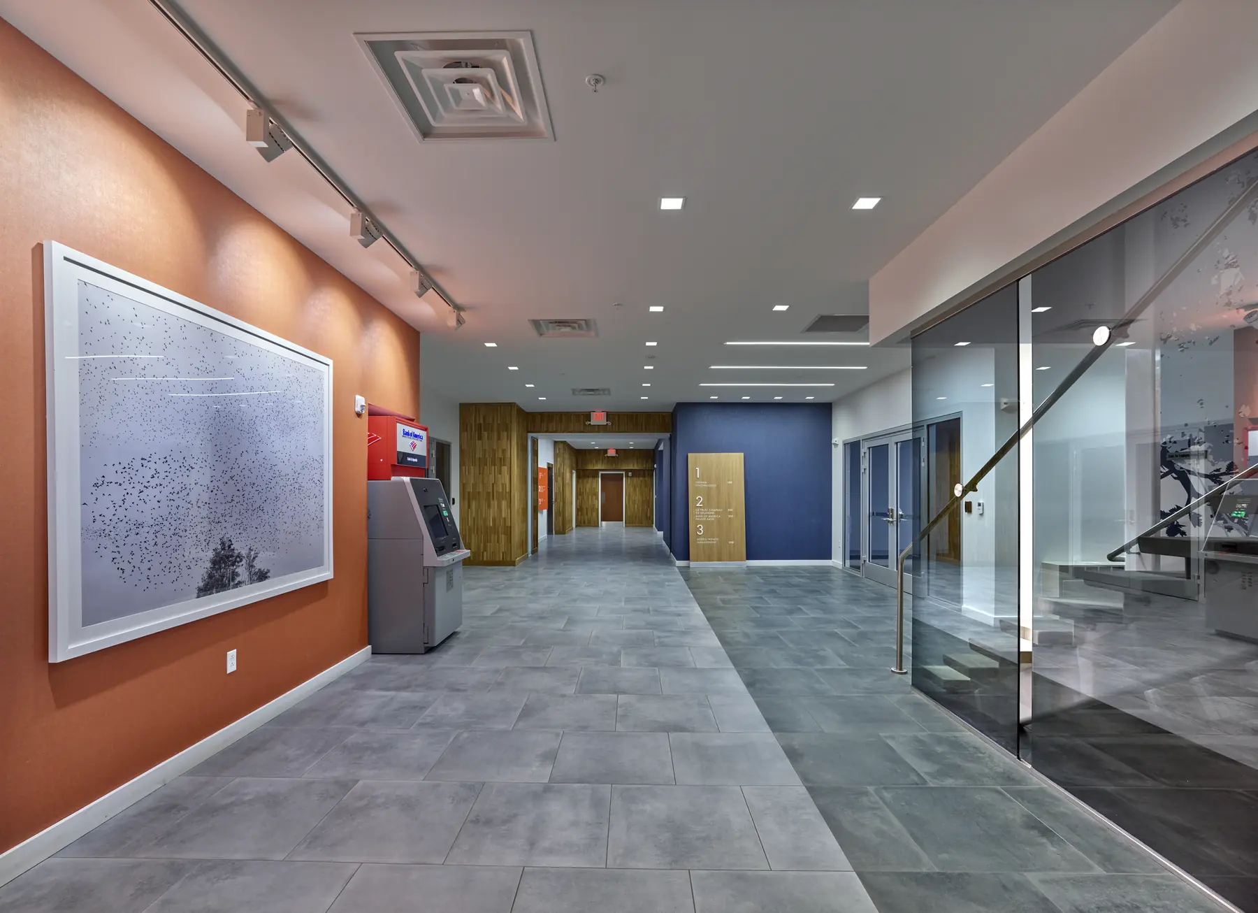Modern office lobby hallway with orange accent wall, artwork, Bank of America ATM, and glass staircase enclosure