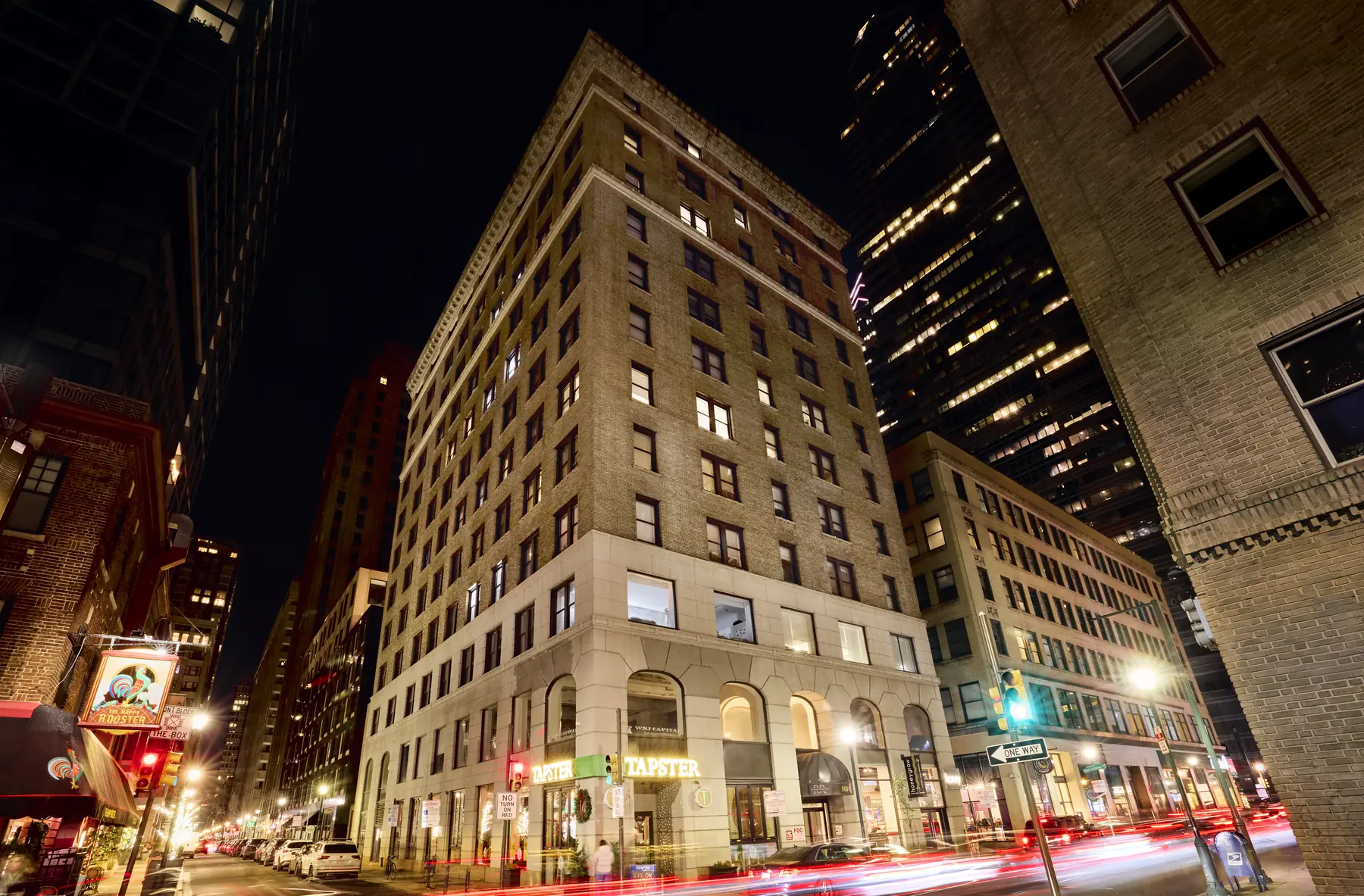 Historic brick office building with Tapster storefront illuminated at night amid downtown high-rises and city traffic.