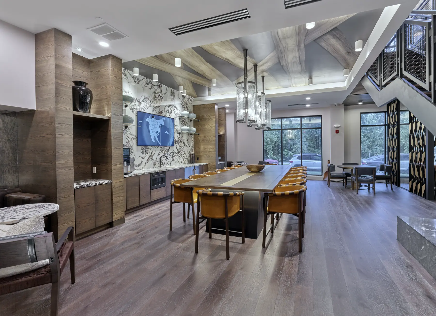 Communal lounge with long dining table, wall-mounted TV, marble accents, and staircase to upper level.