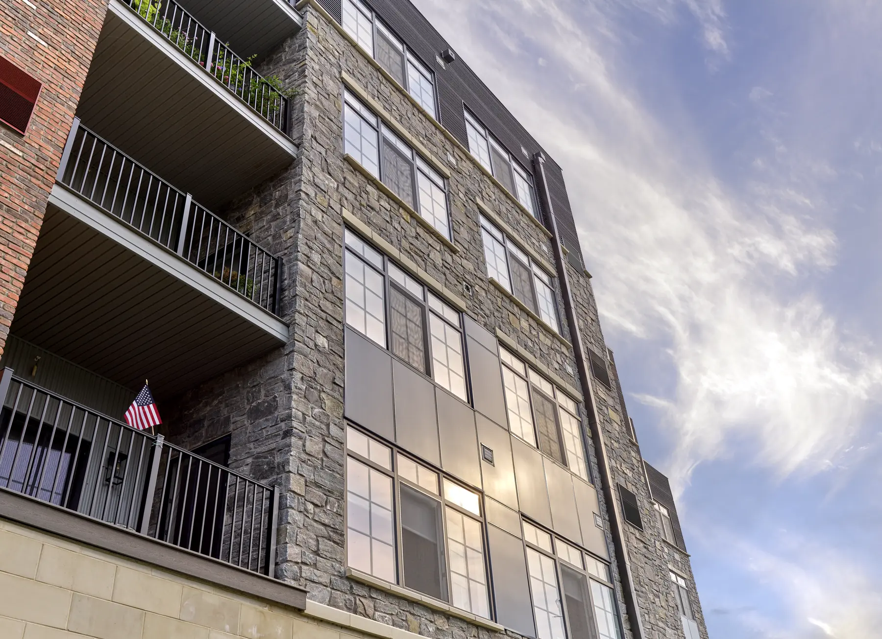Multi-story stone and brick apartment building with private balconies and an American flag against a blue sky
