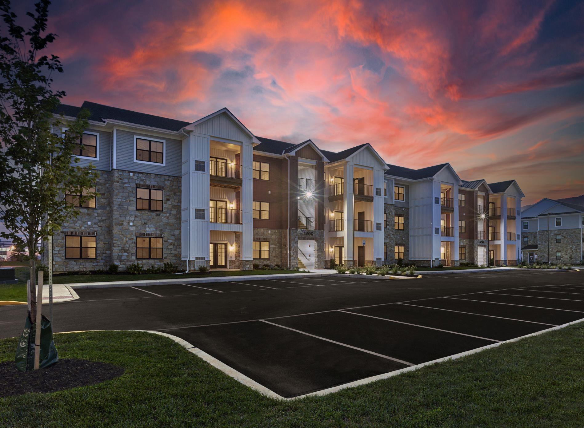 Three-story apartment building with stone and siding exterior, balconies, and a lit parking lot at sunset.