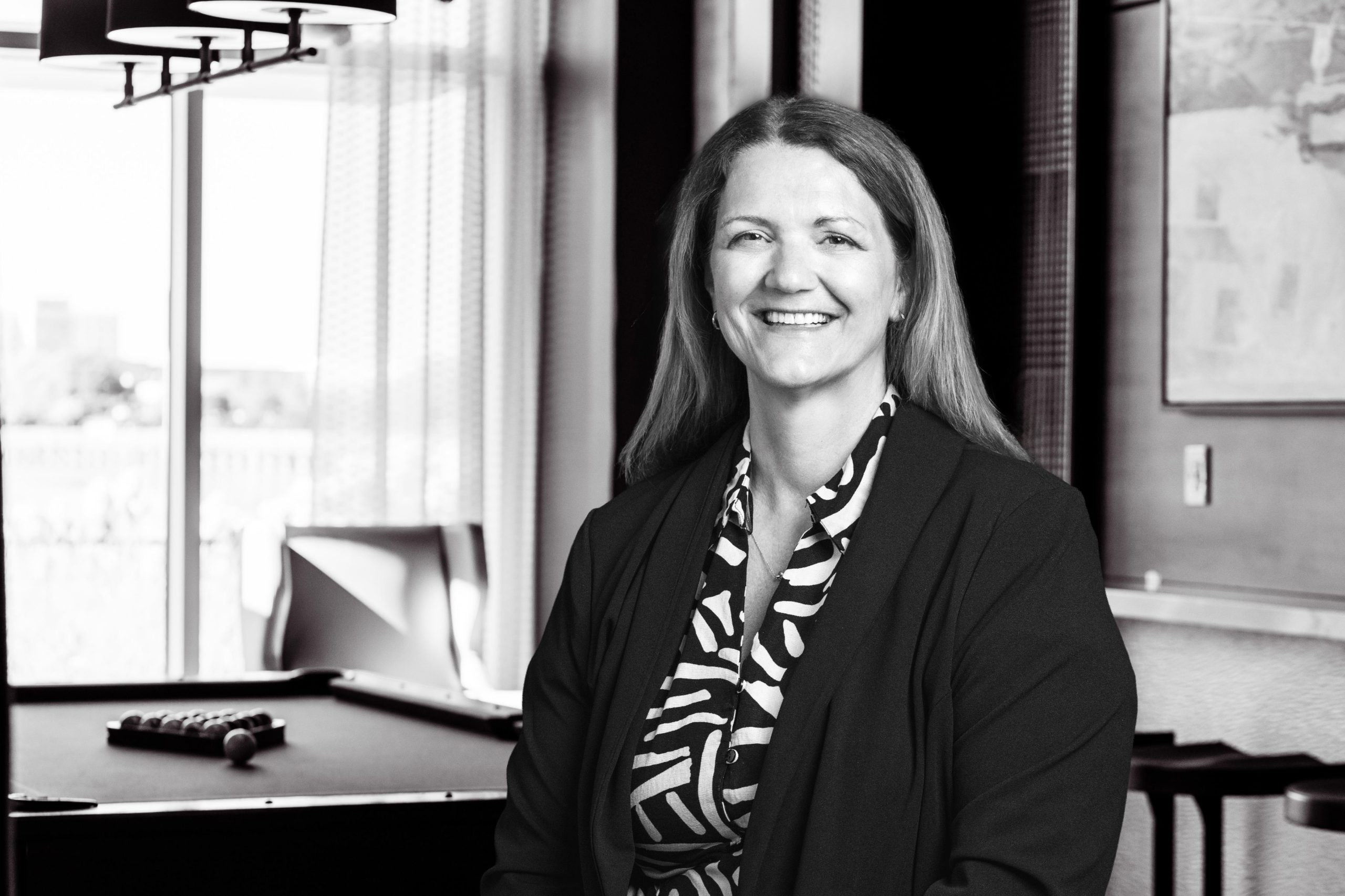 Smiling woman in a black blazer and patterned blouse stands in a lounge area with a pool table nearby.