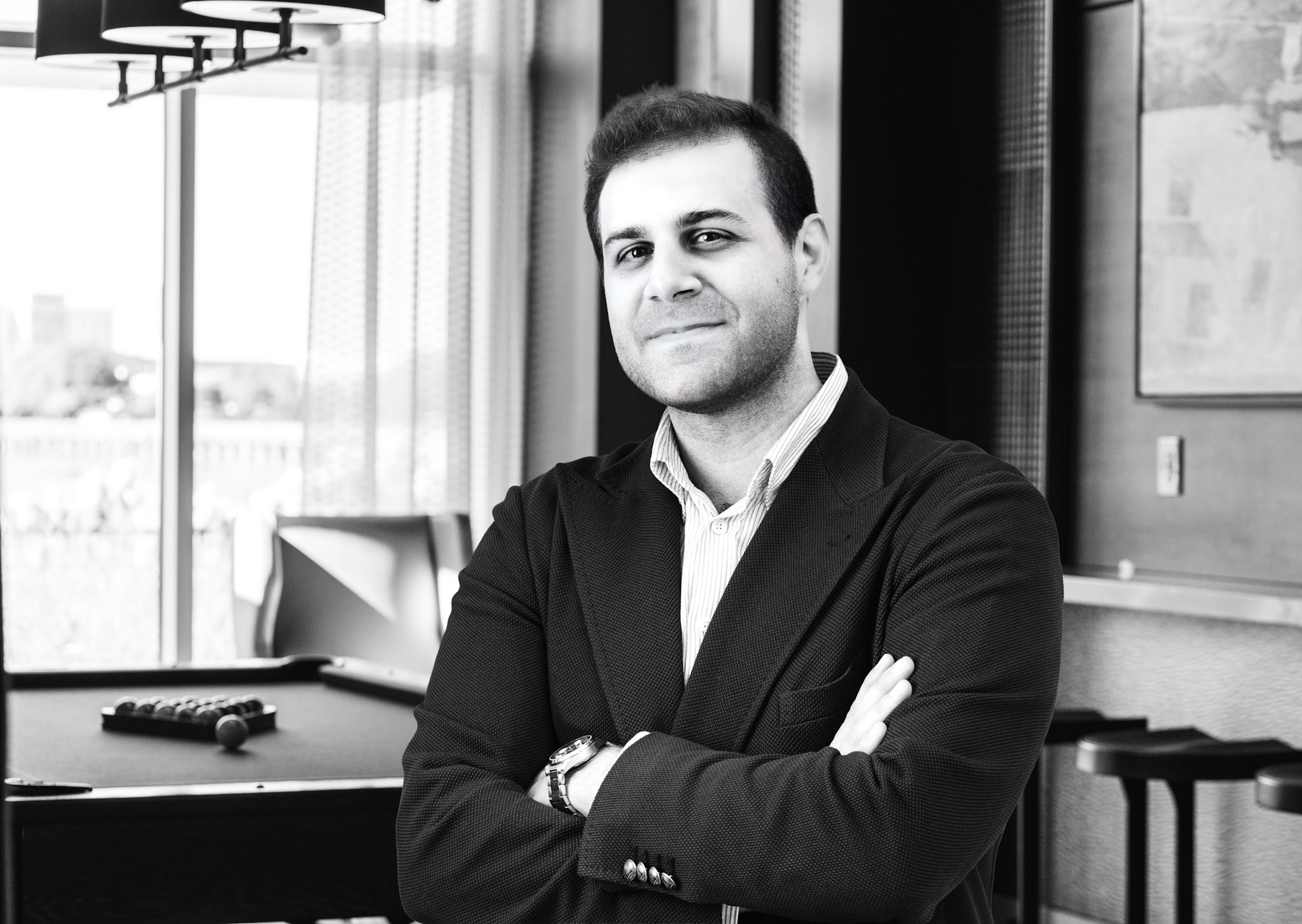 A man in a blazer stands with arms crossed in a lounge area with a pool table and city view.