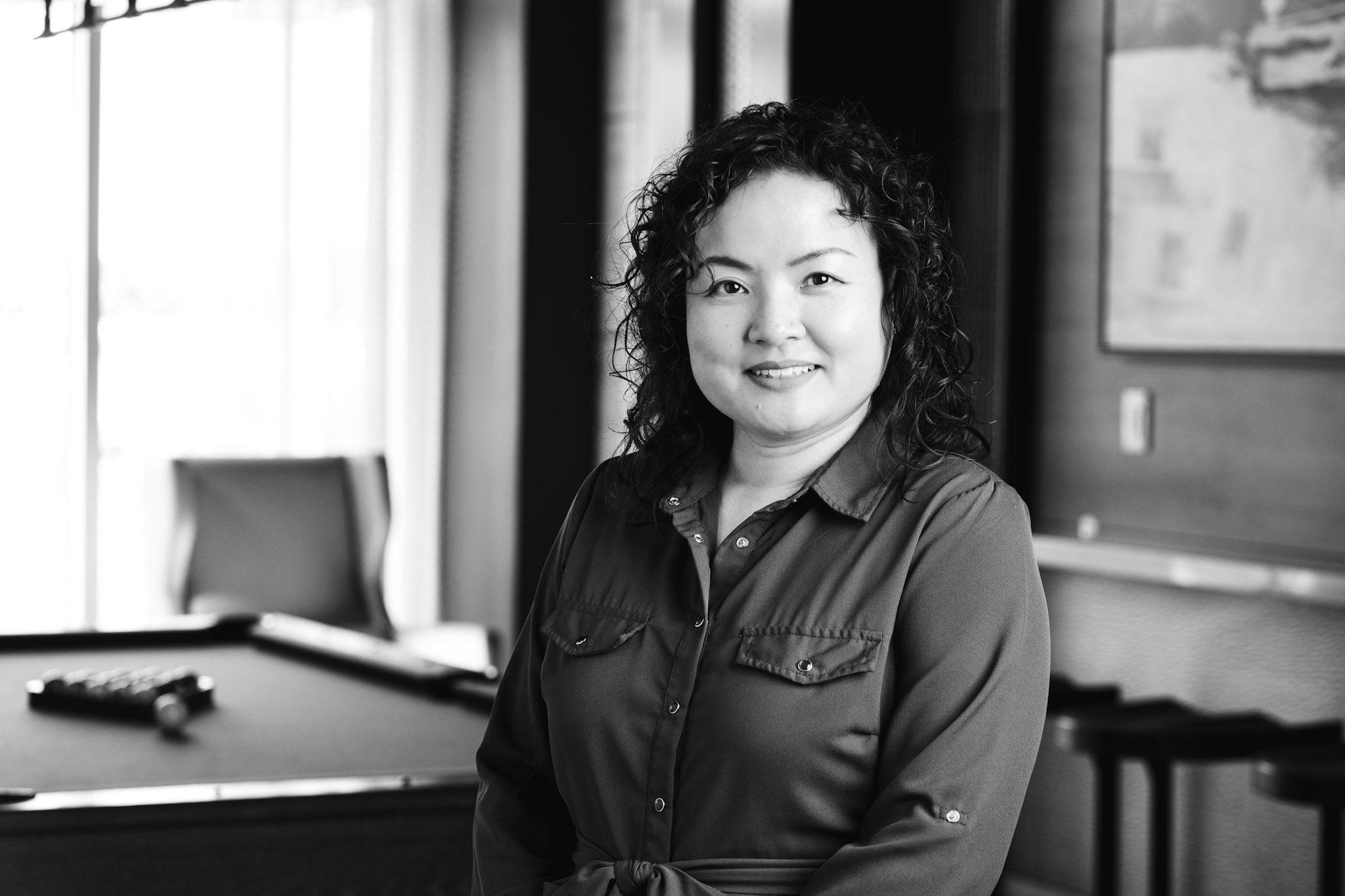 Woman with curly hair smiling in a black button-up shirt in an office setting with a pool table nearby.