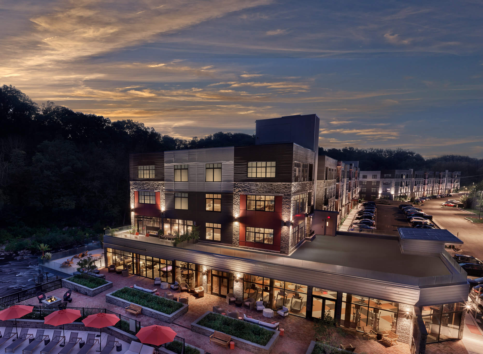 Modern apartment building with lit windows, rooftop patio, and pool lounge area at dusk near a wooded riverside setting