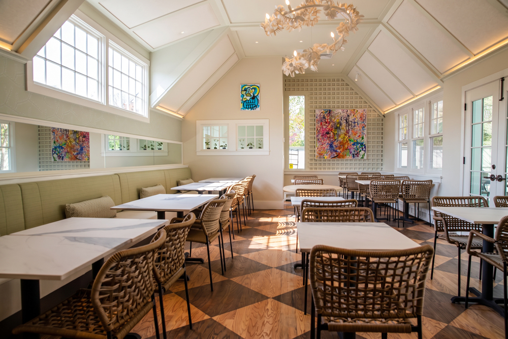Bright dining area with wicker chairs, marble-top tables, and a checkered wood floor under a vaulted ceiling