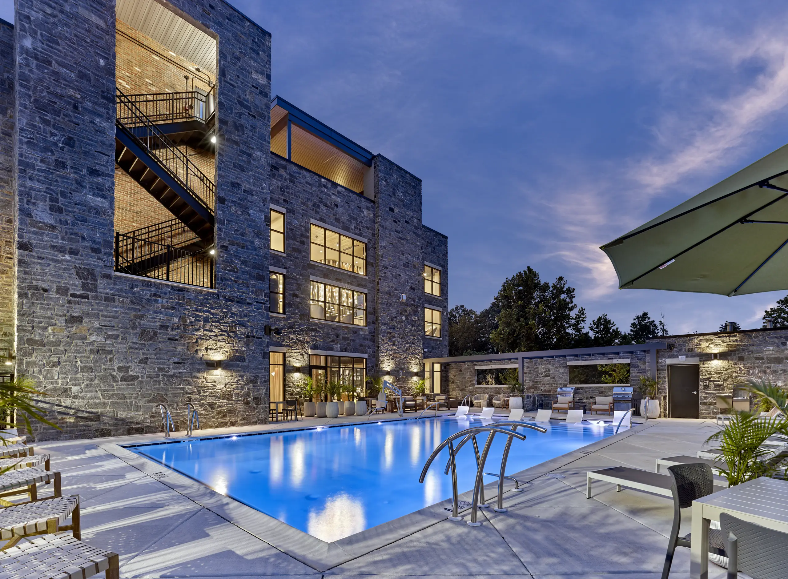 Twilight view of a stone apartment building with a rectangular pool, lounge seating, and outdoor grilling area at The Falls