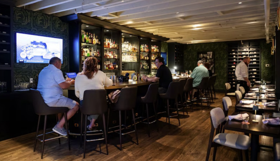 Patrons sit at a well-stocked bar while a waiter attends nearby tables in a dimly lit restaurant
